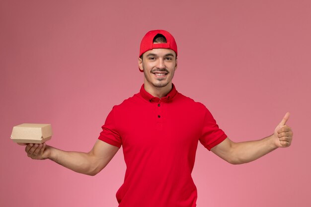 Mensajero masculino de vista frontal en uniforme rojo y capa sosteniendo pequeño paquete de entrega sonriendo sobre el fondo rosa.