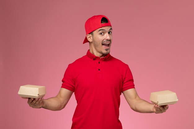 Mensajero masculino de vista frontal en uniforme rojo y capa sosteniendo pequeños paquetes de entrega en el fondo rosa.