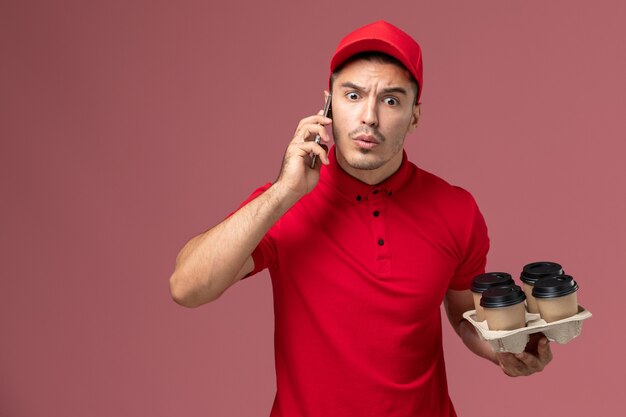 Mensajero masculino de vista frontal en uniforme rojo y capa sosteniendo tazas de café de entrega y hablando por teléfono en la pared rosa