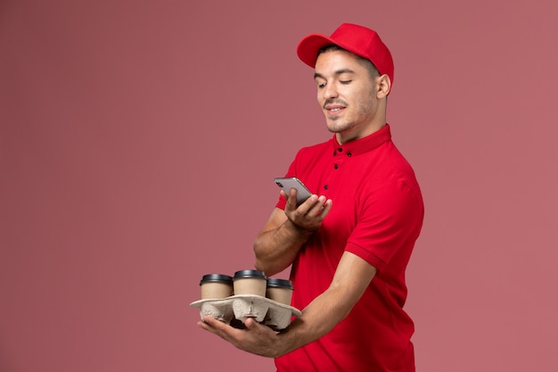 Mensajero masculino de vista frontal en uniforme rojo y capa sosteniendo tazas de café de entrega tomando una foto de ellos en la pared rosa