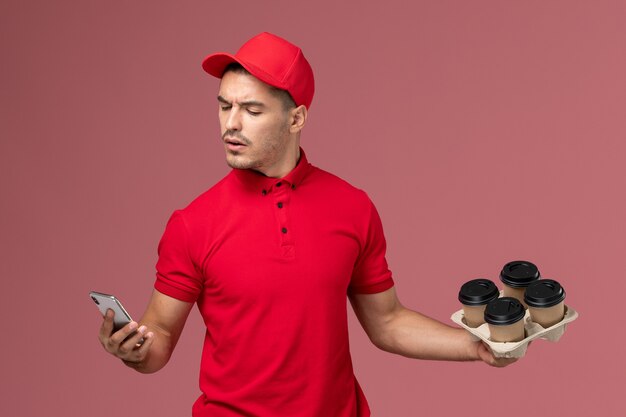 Mensajero masculino de vista frontal en uniforme rojo y capa sosteniendo tazas de café de entrega y usando su teléfono en la pared rosa