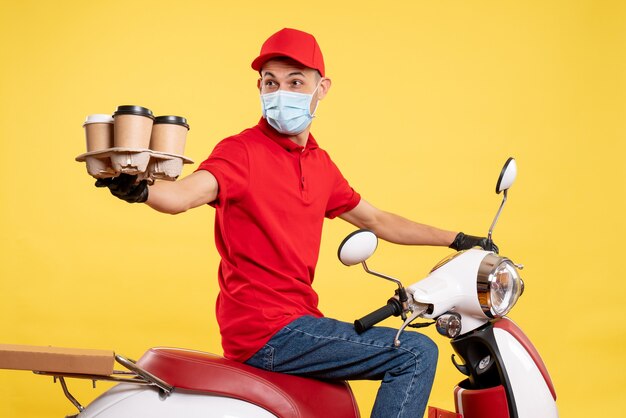 Mensajero masculino de vista frontal en uniforme rojo y máscara con café en el trabajo de color amarillo entrega pandémica covid- trabajo servicio de alimentos