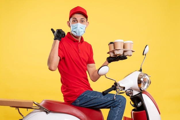 Mensajero masculino de vista frontal en uniforme rojo y máscara con café en el trabajo de color amarillo entrega pandémica covid- trabajo servicio de alimentos