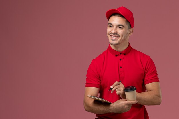 Mensajero masculino de la vista frontal en uniforme rojo que sostiene la taza de café de entrega y el bloc de notas escribiendo notas en el trabajo masculino del trabajador de escritorio rosa