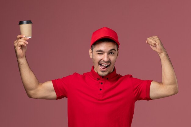 Mensajero masculino de vista frontal en uniforme rojo sosteniendo la taza de café de entrega y flexionando un guiño en el trabajador de la pared rosa