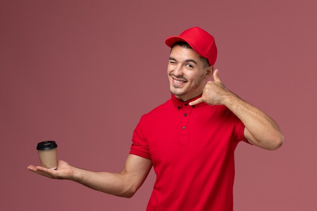 Mensajero masculino de vista frontal en uniforme rojo sosteniendo la taza de café de entrega guiñando un ojo en la pared rosa trabajo masculino