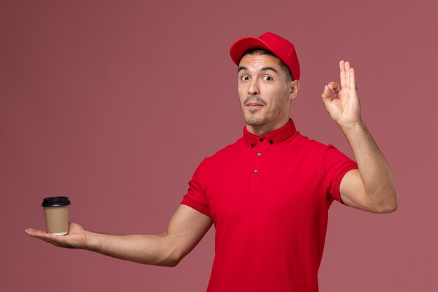 Mensajero masculino de vista frontal en uniforme rojo sosteniendo la taza de café de entrega mostrando bien firmar en la pared rosa trabajo trabajador masculino