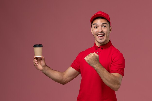 Mensajero masculino de vista frontal en uniforme rojo sosteniendo la taza de café de entrega y regocijándose en la pared rosa trabajador masculino