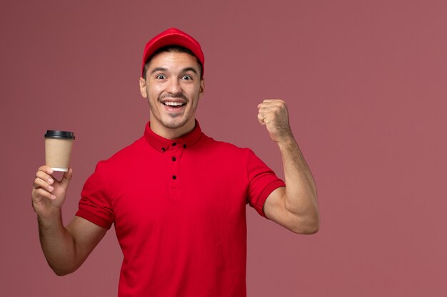 Mensajero masculino de vista frontal en uniforme rojo sosteniendo la taza de café de entrega y regocijándose en la pared rosa trabajador masculino