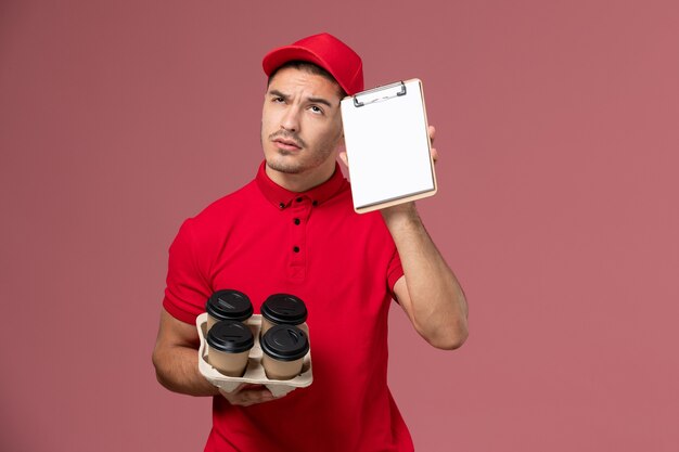 Mensajero masculino de vista frontal en uniforme rojo sosteniendo tazas de café de entrega con bloc de notas pensando en la pared rosa