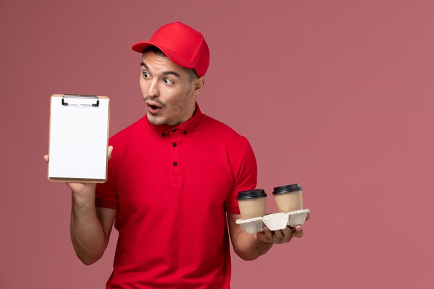 Mensajero masculino de vista frontal en uniforme rojo sosteniendo tazas de café de entrega marrón y bloc de notas en el trabajo de trabajador de pared rosa claro macho