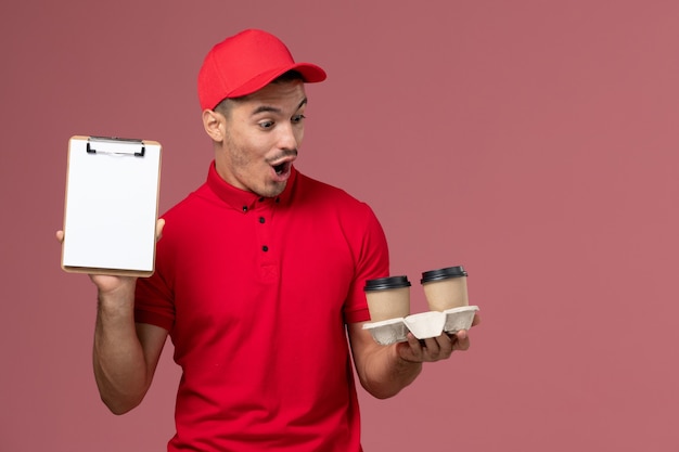 Mensajero masculino de vista frontal en uniforme rojo sosteniendo tazas de café de entrega marrón y bloc de notas en el trabajo de trabajador de pared rosa claro macho