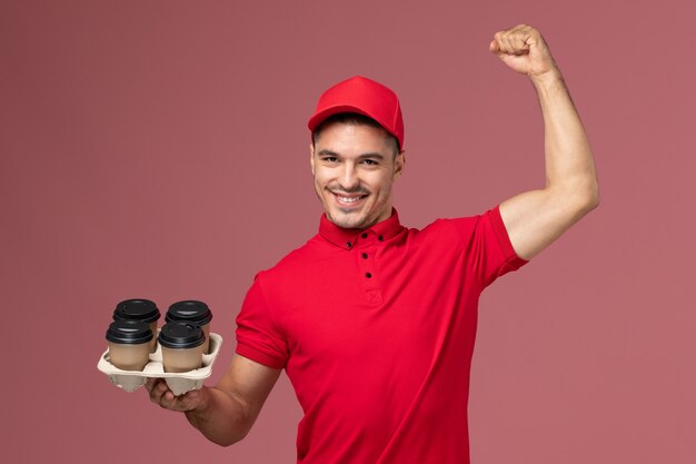 Mensajero masculino de vista frontal en uniforme rojo sosteniendo tazas de café de entrega marrón flexionando sobre trabajador de pared rosa claro