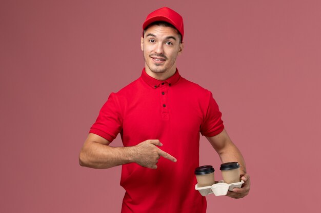 Mensajero masculino de vista frontal en uniforme rojo sosteniendo tazas de café de entrega marrón con una leve sonrisa en la pared rosa macho