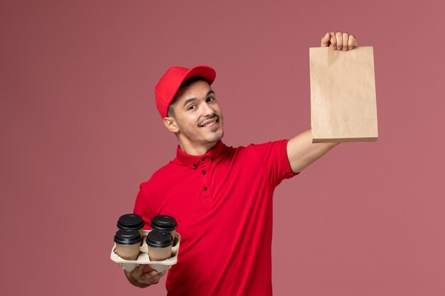 Mensajero masculino de vista frontal en uniforme rojo sosteniendo tazas de café de entrega marrón con paquete de comida en uniforme de trabajador de trabajo de entrega de servicio de escritorio rosa