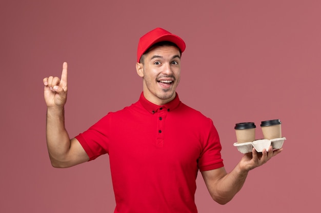 Mensajero masculino de vista frontal en uniforme rojo sosteniendo tazas de café de entrega marrón en la pared de color rosa claro macho