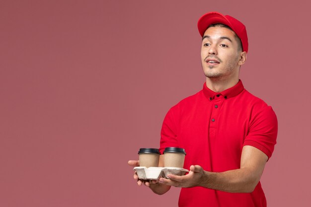 Mensajero masculino de vista frontal en uniforme rojo sosteniendo tazas de café de entrega marrón en la pared rosa uniforme de trabajador de entrega de servicio