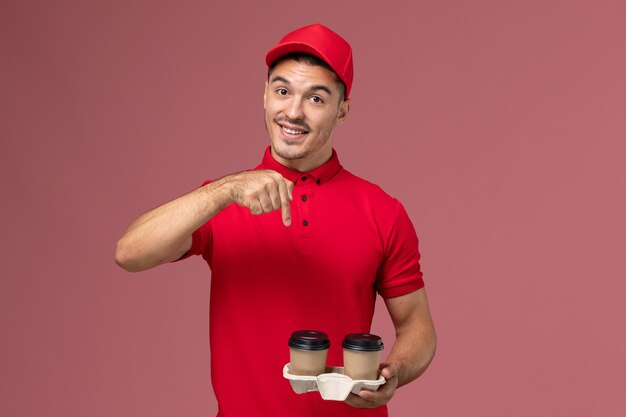 Mensajero masculino de vista frontal en uniforme rojo sosteniendo tazas de café de entrega marrón con sonrisa en el trabajo uniforme de trabajador de entrega de servicio de pared rosa
