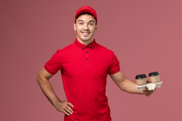 Mensajero masculino de vista frontal en uniforme rojo sosteniendo tazas de café de entrega marrón con sonrisa en el trabajo uniforme de trabajador de entrega de servicio de pared rosa