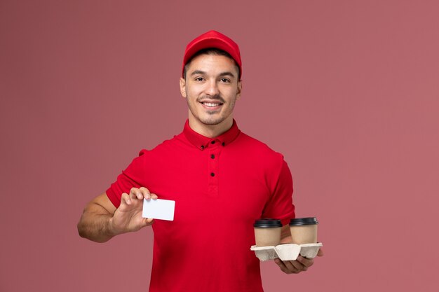 Mensajero masculino de vista frontal en uniforme rojo sosteniendo tazas de café de entrega marrón con tarjeta blanca en la pared rosa claro