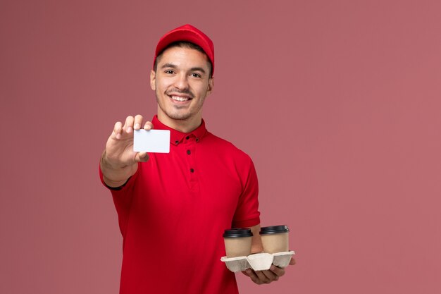 Mensajero masculino de vista frontal en uniforme rojo sosteniendo tazas de café de entrega marrón con tarjeta blanca en la pared rosa claro