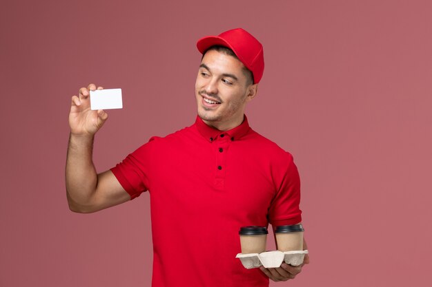 Mensajero masculino de vista frontal en uniforme rojo sosteniendo tazas de café de entrega marrón con tarjeta blanca en la pared rosa claro