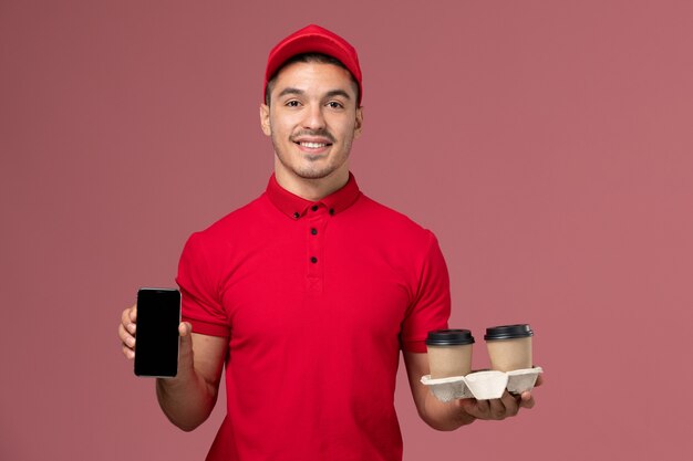 Mensajero masculino de vista frontal en uniforme rojo sosteniendo tazas de café de entrega marrón y teléfono en el macho de trabajo de trabajador de pared rosa claro