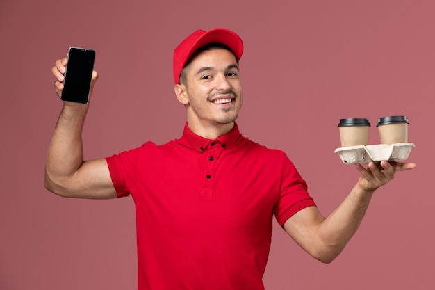 Mensajero masculino de vista frontal en uniforme rojo sosteniendo tazas de café de entrega marrón y teléfono en la pared rosa claro