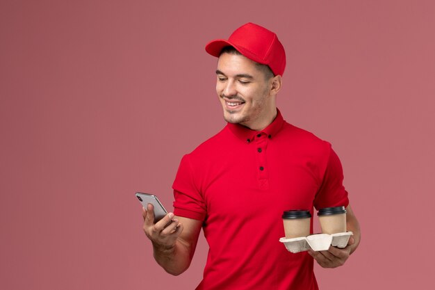 Mensajero masculino de vista frontal en uniforme rojo sosteniendo tazas de café de entrega marrón y teléfono en trabajador de pared rosa claro