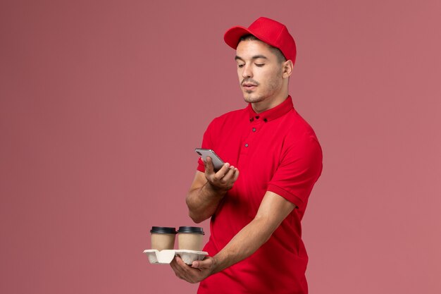 Mensajero masculino de vista frontal en uniforme rojo sosteniendo tazas de café de entrega marrón tomando fotos de ellos en la pared rosa