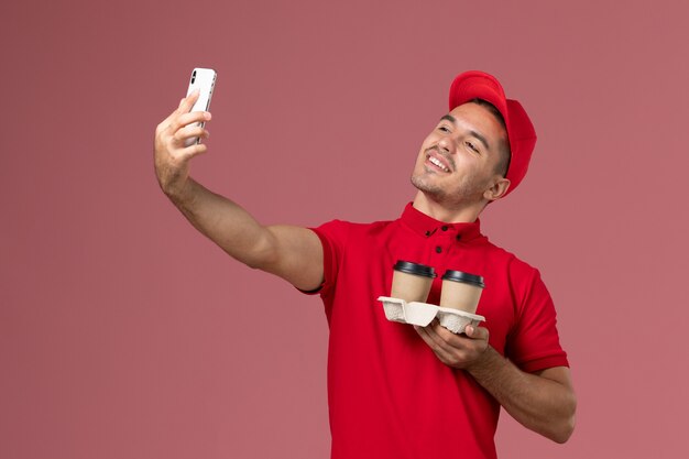 Mensajero masculino de vista frontal en uniforme rojo sosteniendo tazas de café de entrega marrón tomando un selfie con ellos en la pared rosa