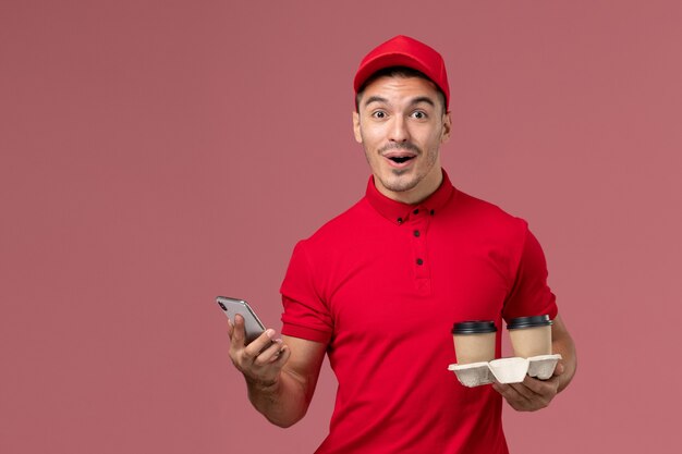 Mensajero masculino de vista frontal en uniforme rojo sosteniendo tazas de café de entrega marrón y usando un teléfono en la pared rosa claro