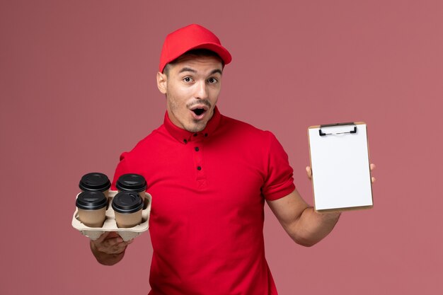 Mensajero masculino de vista frontal en uniforme rojo sosteniendo tazas de café de entrega con un pequeño bloc de notas en la pared rosa