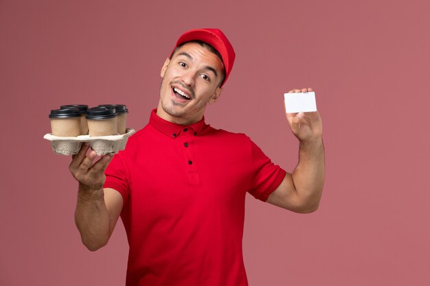 Mensajero masculino de vista frontal en uniforme rojo sosteniendo tazas de café de entrega con tarjeta en el uniforme de trabajador de entrega de trabajo de servicio de pared rosa