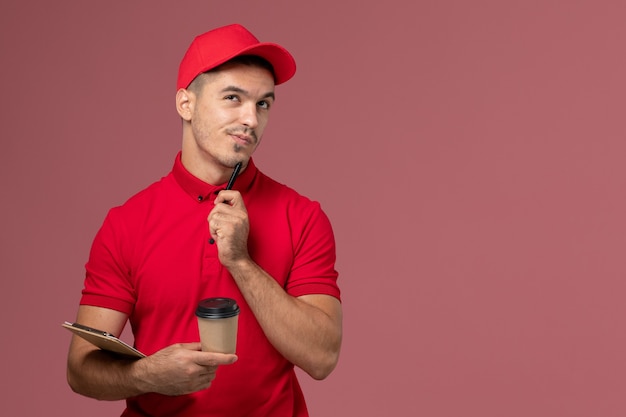Mensajero masculino de vista frontal en uniforme rojo con taza de café de entrega y bloc de notas pensando en la pared rosa