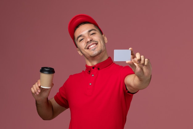 Mensajero masculino de vista frontal en uniforme rojo con taza de café de entrega y tarjeta en el trabajador de escritorio rosa