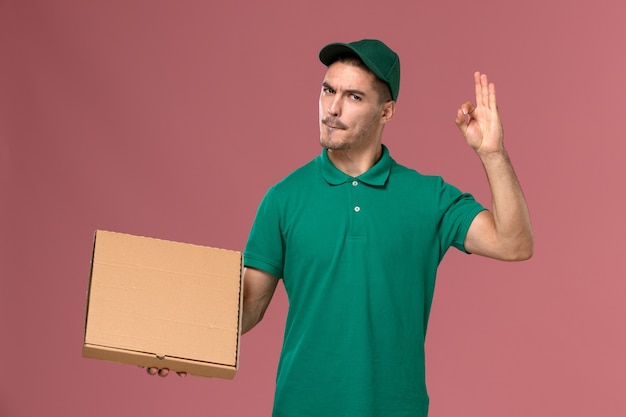 Mensajero masculino de vista frontal en uniforme verde con caja de comida en el fondo rosa