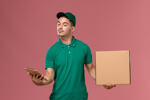 Mensajero masculino de vista frontal en uniforme verde con caja de comida junto con el bloc de notas en el escritorio rosa