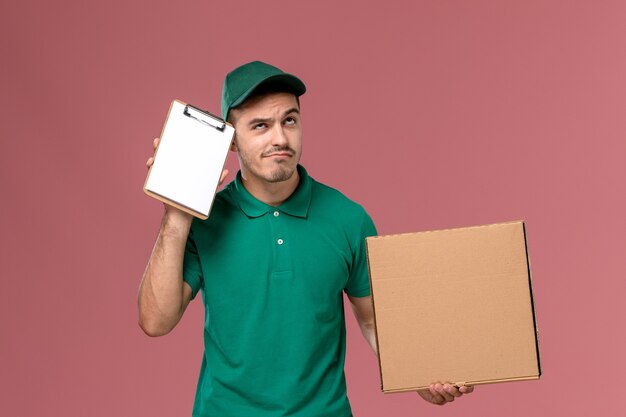 Mensajero masculino de vista frontal en uniforme verde con caja de comida junto con el bloc de notas pensando en fondo rosa claro