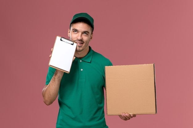 Mensajero masculino de vista frontal en uniforme verde con caja de comida junto con un pequeño bloc de notas sobre fondo rosa claro