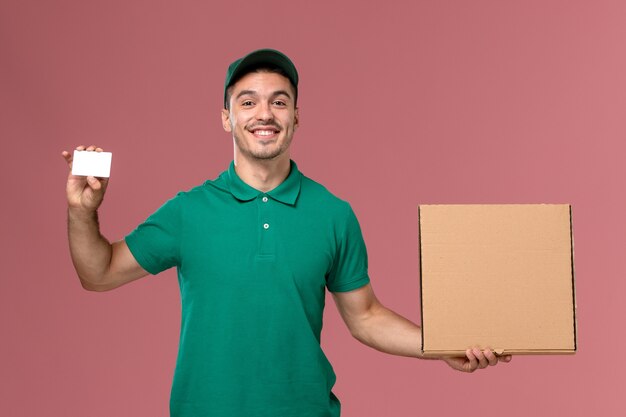 Mensajero masculino de vista frontal en uniforme verde con caja de comida con tarjeta blanca sobre fondo rosa