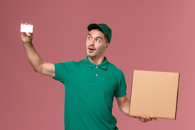 Mensajero masculino de vista frontal en uniforme verde con caja de comida con tarjeta blanca sobre fondo rosa