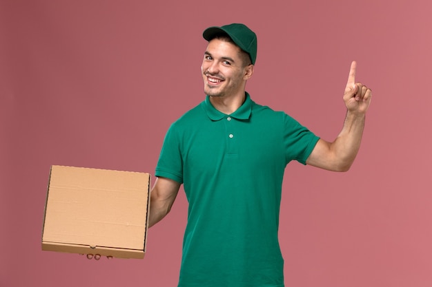 Mensajero masculino de vista frontal en uniforme verde con caja de entrega de alimentos con el dedo levantado sobre fondo rosa