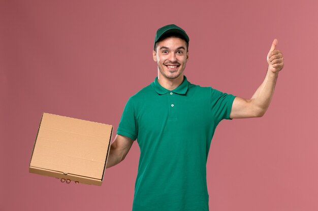 Mensajero masculino de vista frontal en uniforme verde con caja de entrega de alimentos con expresión de deleite en el fondo rosa