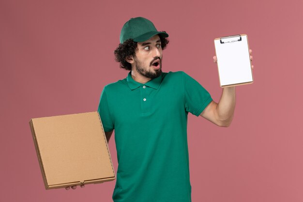 Mensajero masculino de vista frontal en uniforme verde y capa con bloc de notas de caja de comida de entrega en el trabajo de entrega uniforme de servicio de fondo rosa