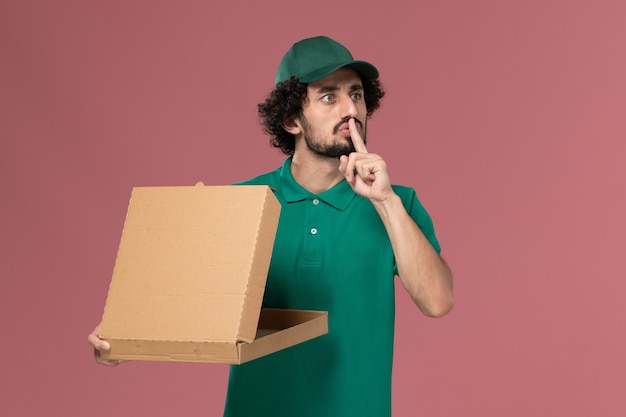 Mensajero masculino de vista frontal en uniforme verde y capa con caja de comida de entrega sobre fondo rosa Servicio de entrega uniforme del trabajador