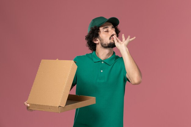 Mensajero masculino de vista frontal en uniforme verde y capa con caja de comida de entrega en el trabajo de entrega uniforme del trabajador de servicio de fondo rosa claro