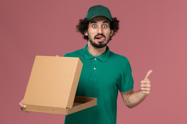 Mensajero masculino de vista frontal en uniforme verde y capa con caja de comida de entrega vacía sobre fondo rosa claro Servicio de entrega uniforme del trabajador
