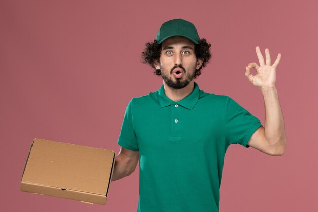 Mensajero masculino de vista frontal en uniforme verde y capa con caja de comida en el fondo rosa servicio trabajador trabajo uniforme entrega