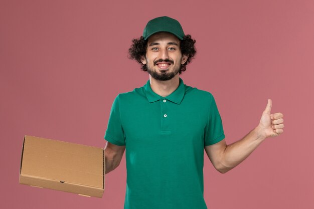 Mensajero masculino de vista frontal en uniforme verde y capa con caja de comida en el trabajo de entrega uniforme del trabajador de servicio de fondo rosa